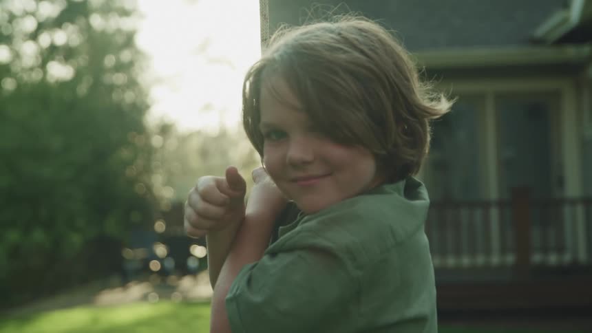 A boy smiles and gives a thumbs up to the camera as he rides a swing in the backyard of a house