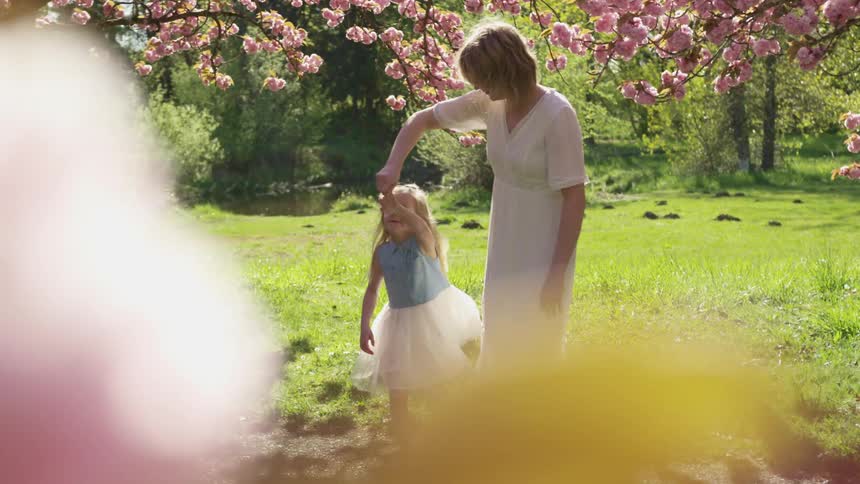 A woman twirls her dancing daughter in a field beneath a blossoming tree on a sunny day