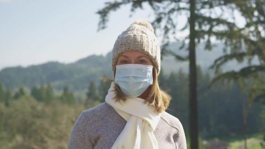 Woman with a grey beanie a white scarf around her neck and a light blue mask faces the camera with eye contact in an open field with mountains in the background