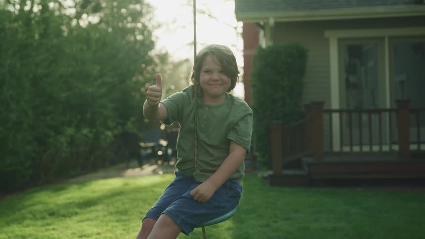 A young boy sitting on a rope swing and smiles while giving a thumbs up