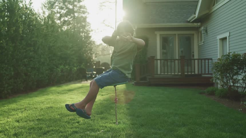 A young boy holds his hand up as he sits on a rope swing in his back yard