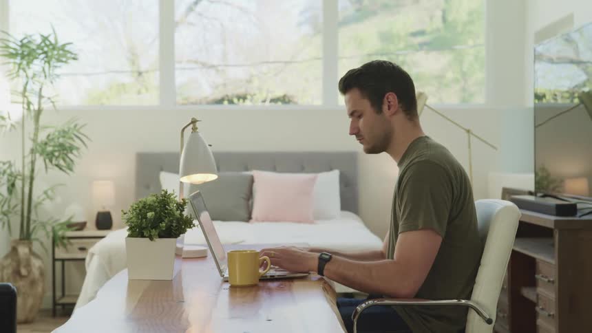 Man with a green t shirt sitting at a desk uses a laptop next to bed in bedroom