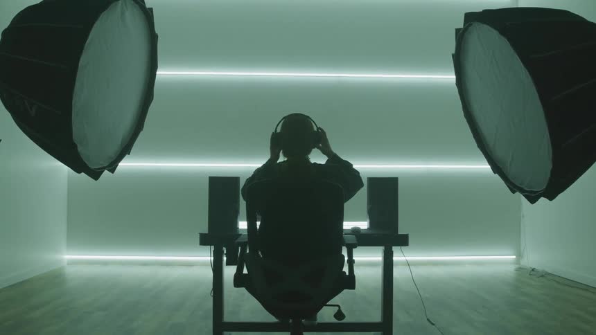 A young woman sitting in a chair at a computer desk slowly puts on a pair of headphones