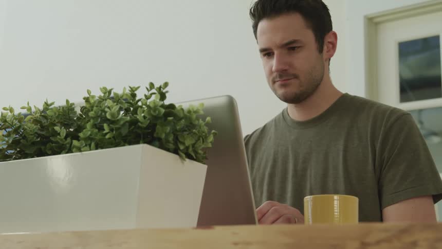 Male sitting at desk smiles slightly while looking at the screen on the laptop in front of him and begins to type on the keyboard