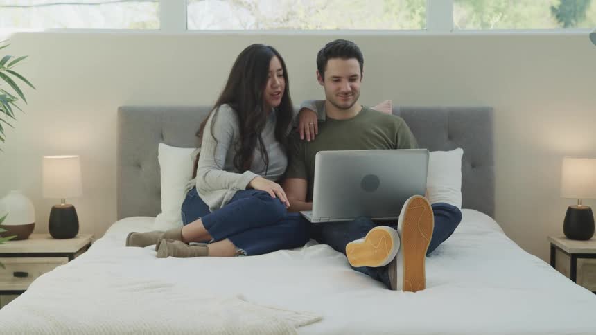 A man sits on a bed with a laptop on his lap as a young woman leans on his shoulder and looks at the screen with him