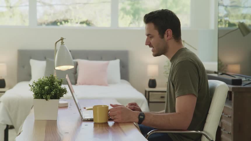 Man talking on a laptop video call on a bedroom while drinking hot beverage