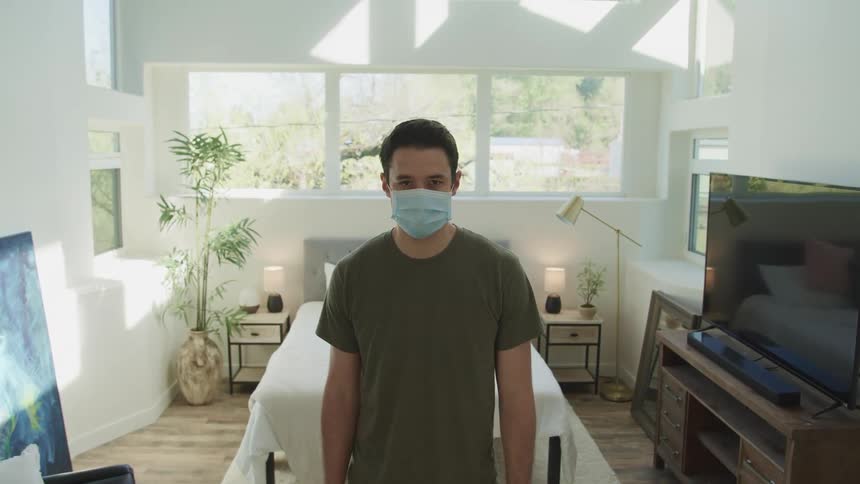 Man with dark green t shirt and a light blue mask raises his head and faces the camera with eye contact in front of a bed next to a television and an oil painting in a room