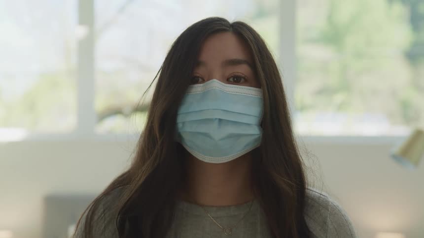 Young woman wearing a blue face mask looks at the camera while in a bedroom at hom