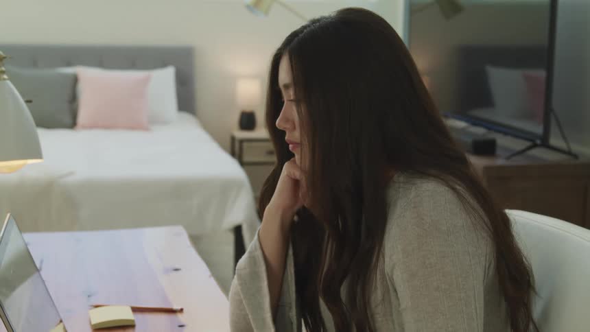 Asian woman with black long hair sitting at a desk looks at a laptop and nods her head in the bedroom
