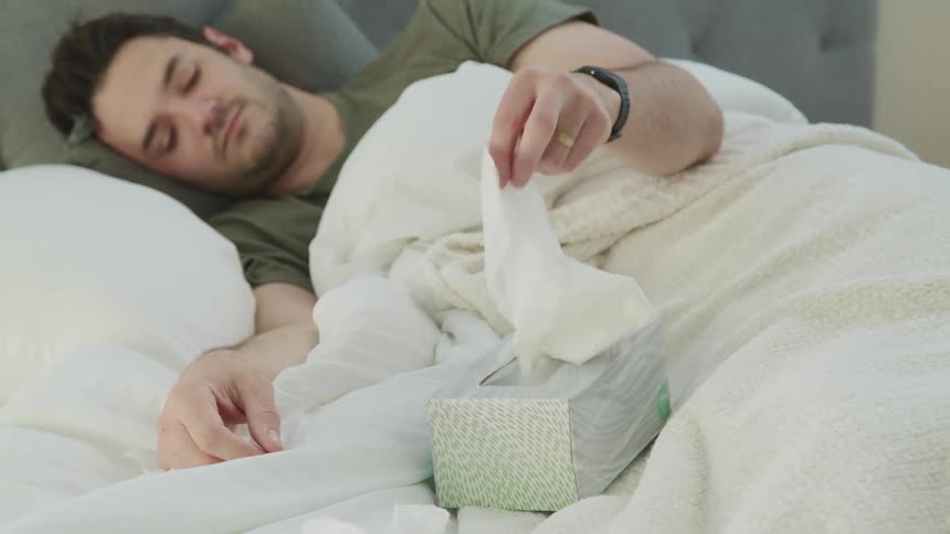 Unshaven male laying on his side in bed while trying to remove a tissue from the tissue box next to him