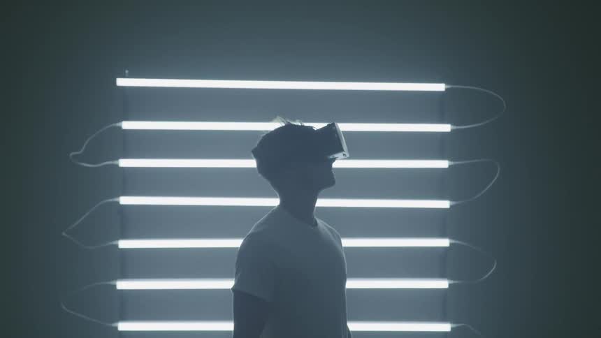 Young man wearing virtual reality goggles stands in front of fluorescent lights in a dimly lit room
