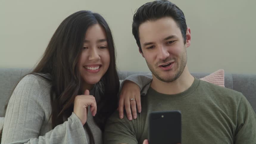 Young couple sitting in a couch wave at someone on a smartphone video call at home