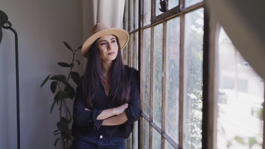 Woman with long black hair a black shirt and a brown hat looks down then looks out the window in a room