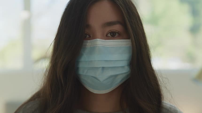 Asian woman with long brown hair and a blue mask faces the camera with eye contact