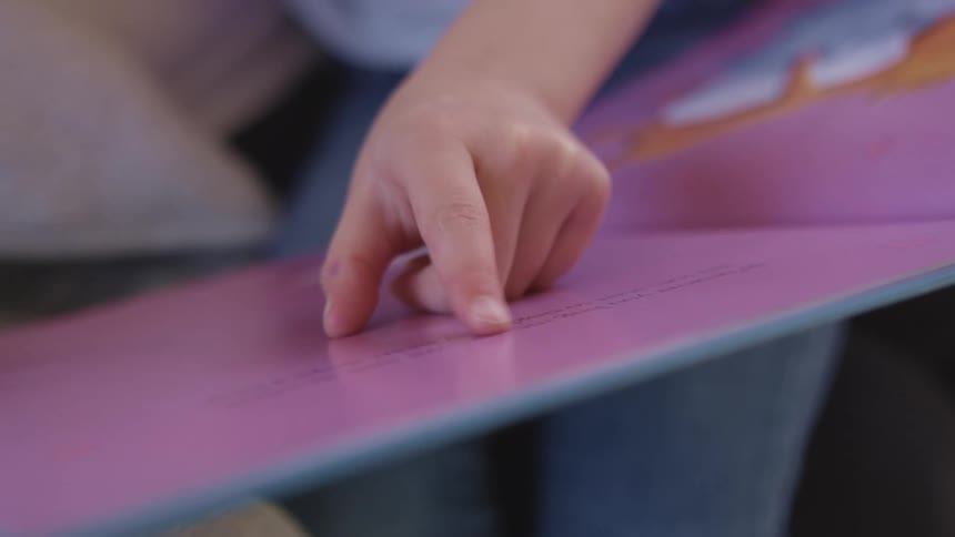 A child points to the words on the page of a story board while a woman sitting next to her holds the book in place
