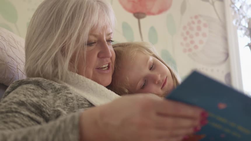 A little girl sits in her grandmothers knee as she reads a book to her