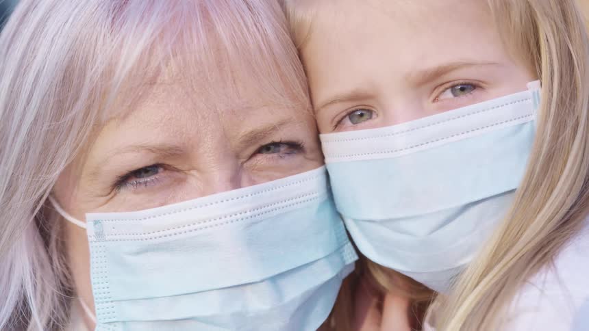Grandmother with grey hair and granddaughter with blonde hair wear light blue masks and cuddle each other