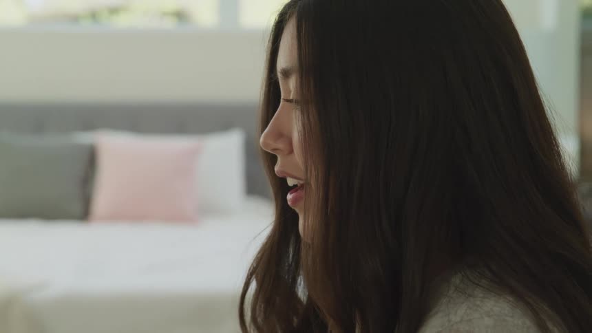 An asian woman sits in a bedroom speaking to someone out of frame