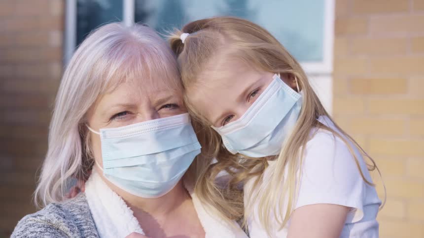 Grandmother with grey hair and granddaughter with blonde hair wear light blue masks and cuddle each other