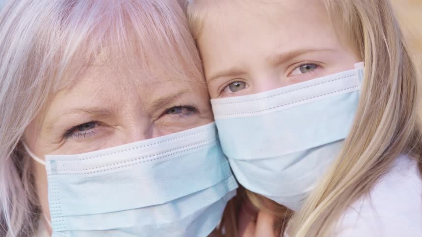 Grandmother with grey hair and granddaughter with blonde hair wear light blue masks and cuddle each other