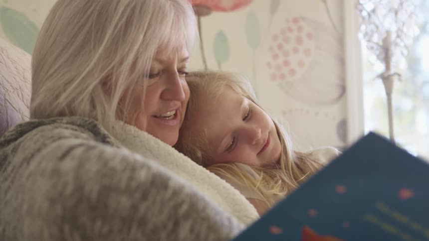 A little girl sits on her grandmas knee and they read a book together