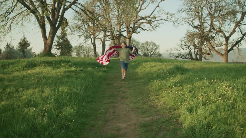 A young boy runs down a dirt path through a field with an american flag draped over his shoulders