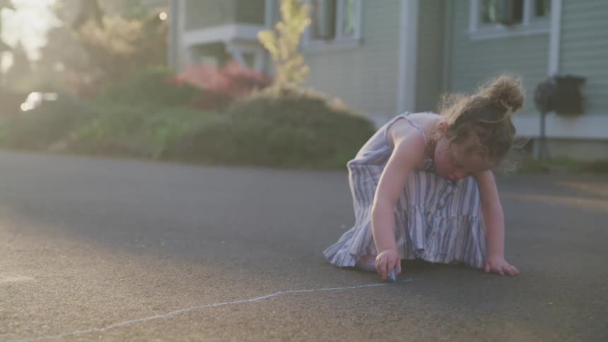 A young girl draws on the road with blue chalk on a sunny day