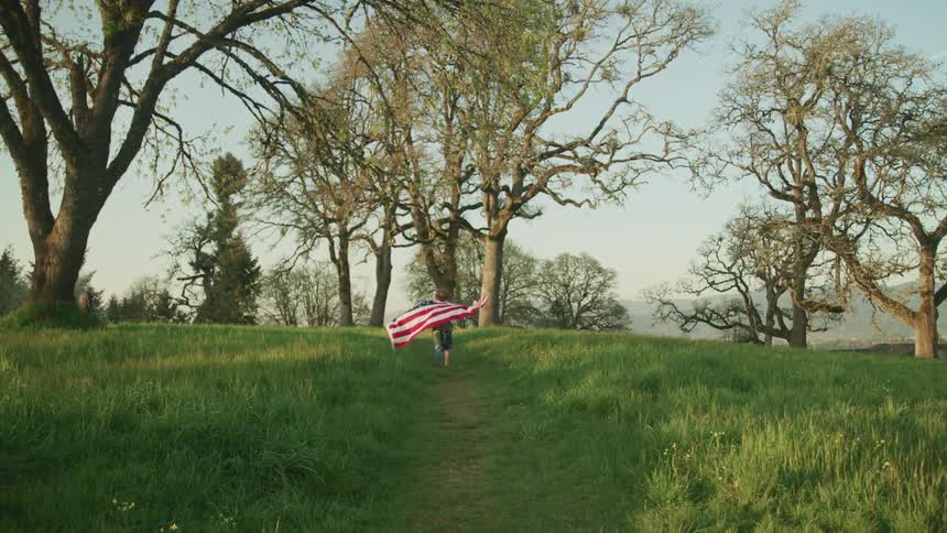 An american flag flaps in the wind as a a child pulls it behind as they run through a field