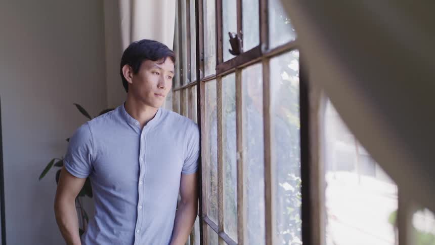 Asian man with a blue shirt looks out the window on a sunny day
