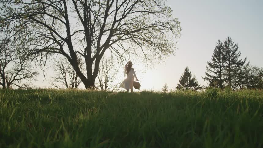 Woman walks across field toward rising sun carrying basket and holding on to her hat
