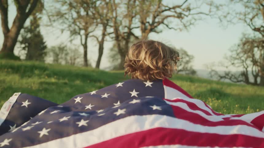 A little boy runs across the grass with an american flag over his shoulders