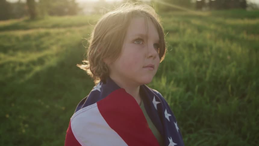 A boy standing in a grassy field draped in an american flag with trees in the background