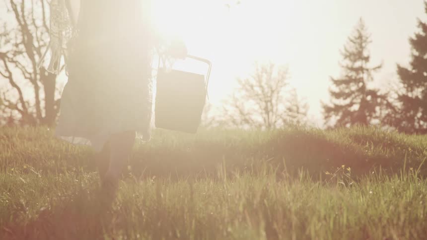 A woman walks through a field in a white dress and carrying a picnic basket