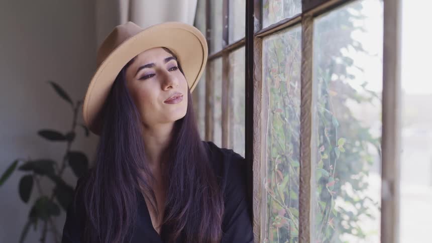 Woman with long black hair and a brown hat looks out the window in a room
