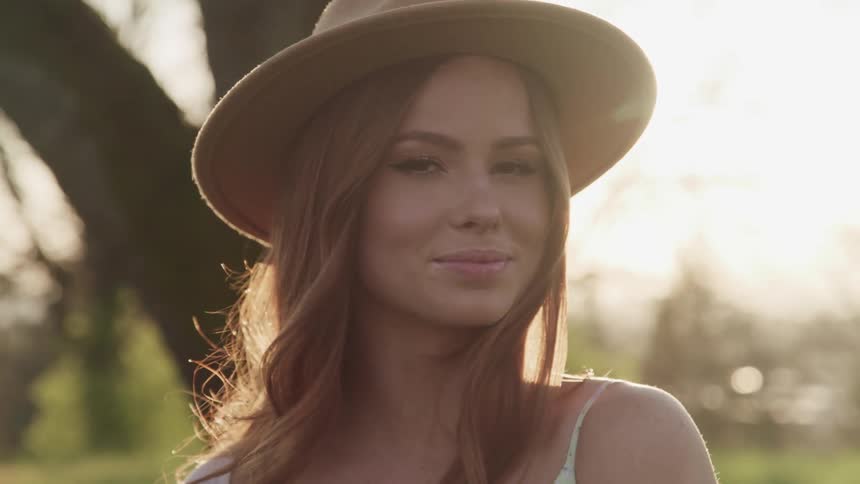Blonde woman with wide hat smiles and faces the camera with eye contact as the sun shines brightly
