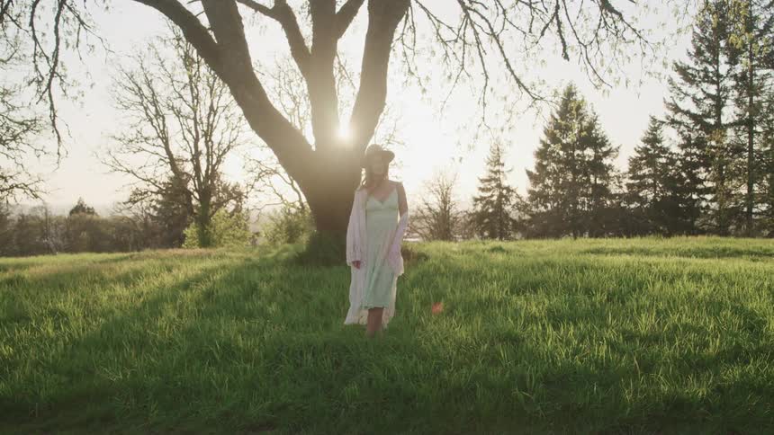 Woman with white dress a hat stands on green grass in front of a tree while the sun shines brightly in the background
