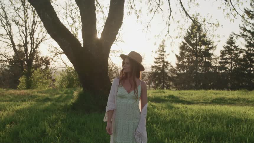 The sun shines between the thick branches of a tree onto a woman standing in a field wearing a sunhat