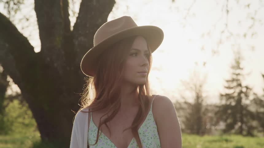 Woman with a brown hat and wavy brown hair looks to the left under a tree as the sun sets in the background