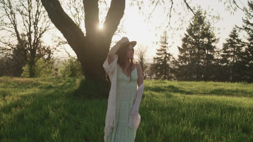 A woman standing outside in a grassy area holds on to her hat