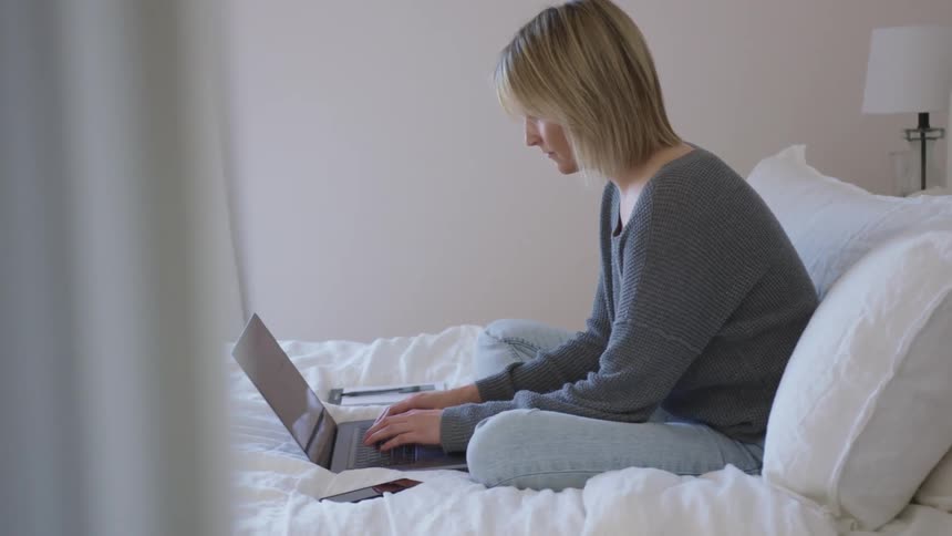 A woman sits on her bed and types on her laptop