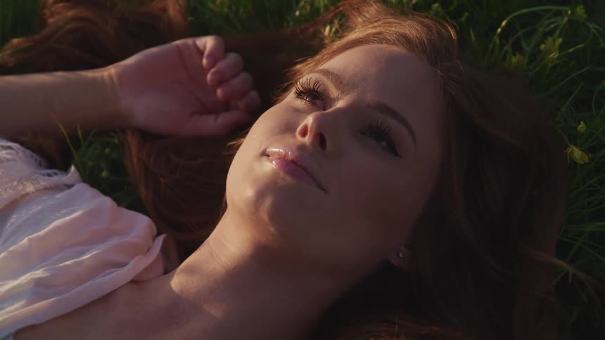 Woman with long brown hair and makeup lies down on green grass at sunset