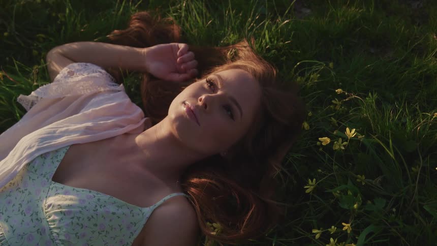 Woman with brown hair and makeup lies down on green grass and relaxes