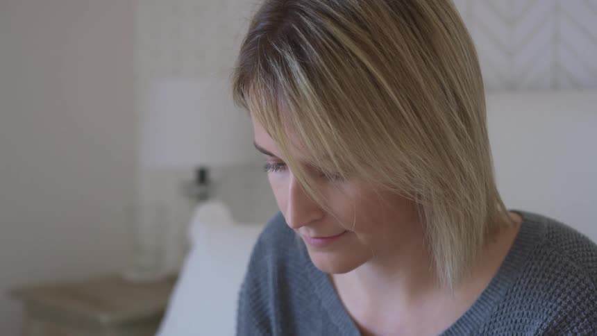Woman with dirty blonde hair and a grey sweater looks down and uses a laptop on a bed in the bedroom