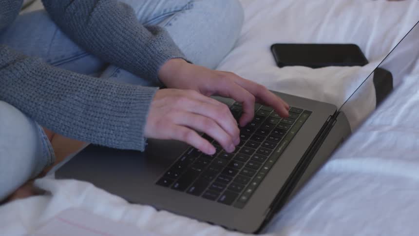 A person sitting on a soft bed types on the keyboard of their metallic gray laptop