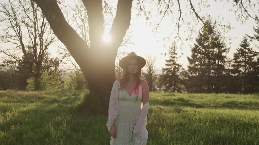 Woman with a white dress a pink shawl and a brown hat stands on green grass and faces the camera with eye contact at sunset