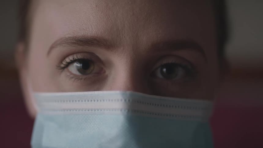 Woman with a ponytail and a light blue mask faces the camera with eye contact