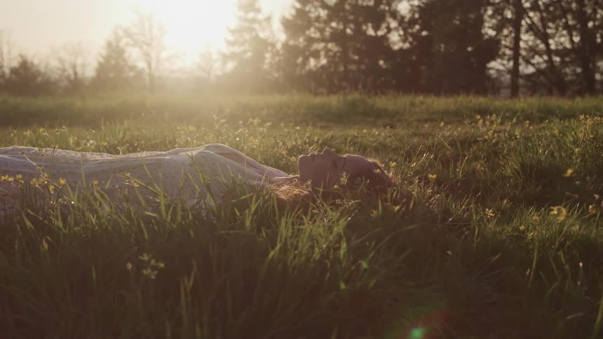 Woman with brown hair lies down on green grass closes her eyes relaxing at sunset