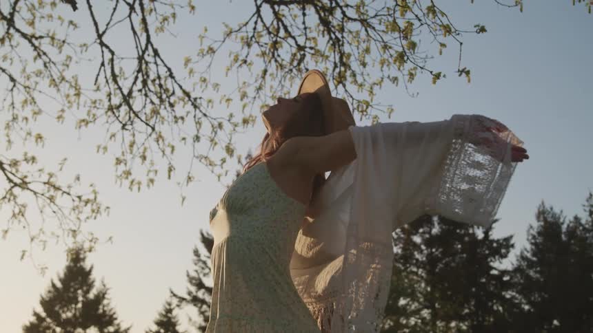 Woman with a brown hat a white dress and a pink shawl looks up and spreads her arms wide under a tree at sunset