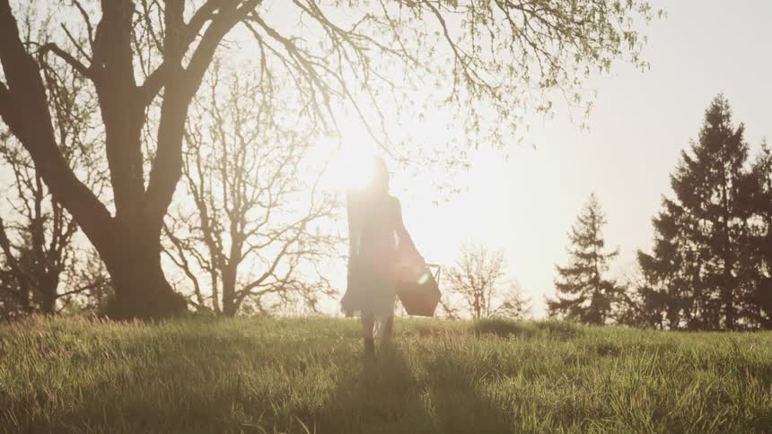 Woman in a white long dress carries a picnic basket on a grass field at sunset