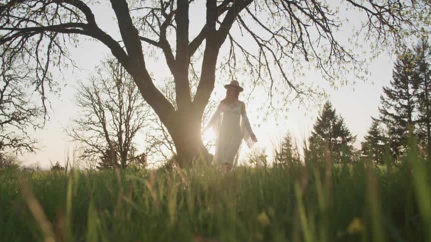 As the sun hangs low in the sky a young woman stands in a field under a tree